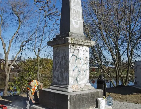 A construction worker works on the Newkirk Monument, a squat, pale grey obelisk, sits on the grass by a path with a blue sky behind